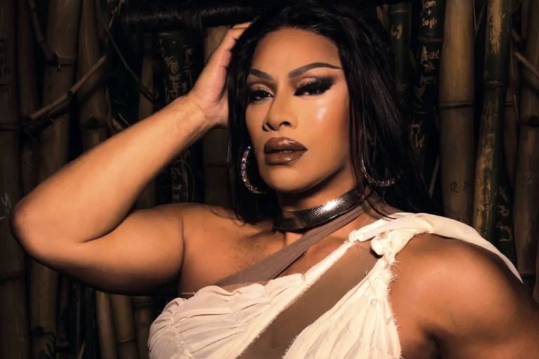 Drag performer Kween Kong posing in full glamour makeup and hoop earrings against a dark bamboo backdrop for a promotional artist portrait photo shoot.